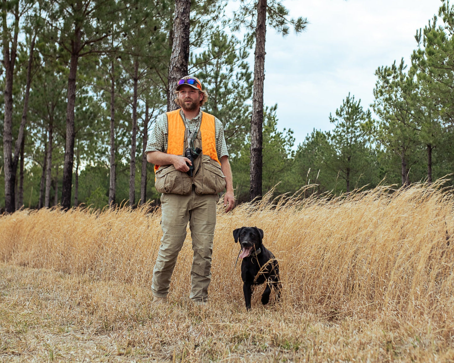 Man in hunting attire with a dog in a field of tall grass and trees.