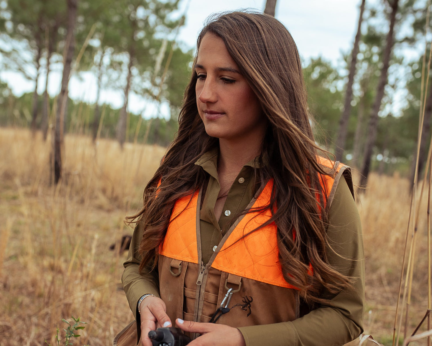 Woman in hunting attire with orange vest and brown jacket in a forest setting