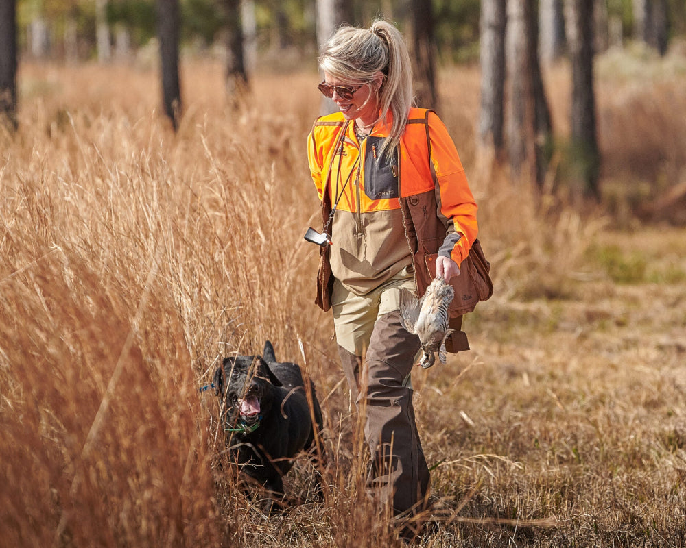 Woman in orange hunting vest walking a black dog through a field with trees in the background